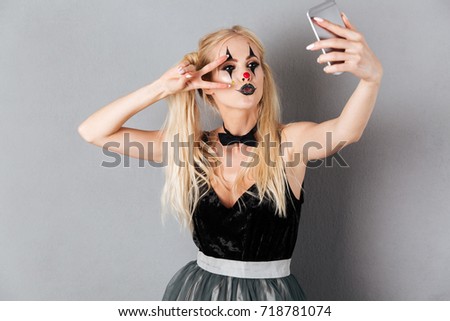 Portrait of an attractive blonde woman in halloween clown make-up taking a selfie with mobile phone isolated over gray background