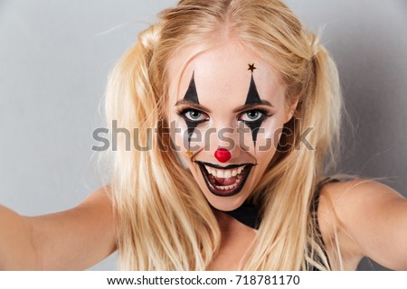 Close up portrait of a pretty smiling blonde woman in bright halloween clown make-up taking a selfie isolated over gray background