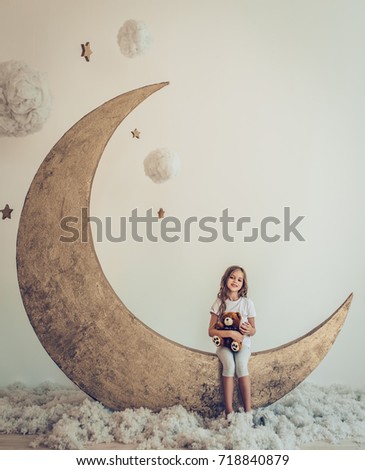 Sweet dreams. Little cute girl with plush toy is sitting on artificial Moon with cotton clouds and stars