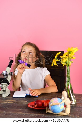 Girl with dreamy face holds purple marker. Schoolgirl at desk near yellow flowers, microscope, globe and fruit. Kid and school supplies on pink background. Back to school and natural beauty concept