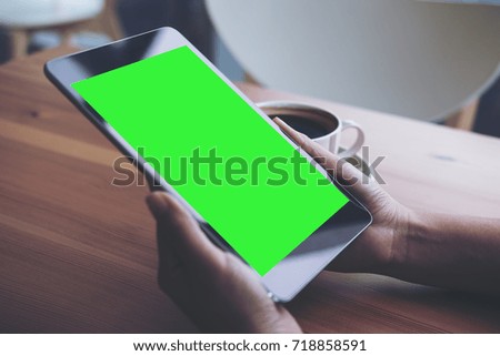 Mockup image of business woman's hands holding black tablet pc with blank green screen and coffee cup on wooden table in cafe background