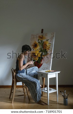 Young female artist at work with brushes, palette, paints draw in oil the picture with sunflowers bouquet, standing on easel. White wall as background. Atmospheric day light.