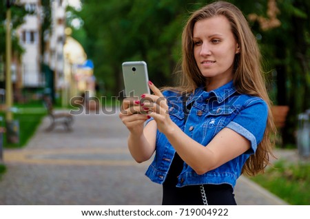 beautiful slim fashionable woman in a black dress and denim blue jacket photographs on a smartphone in a park near the house