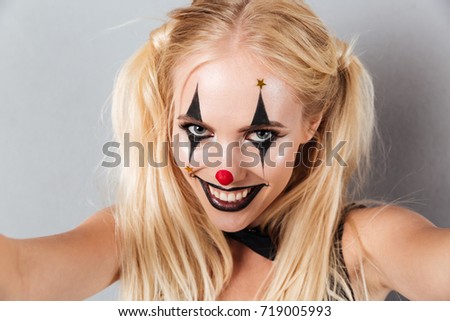 Close up portrait of a cheerful joyful blonde woman in bright halloween clown make-up taking a selfie isolated over gray background