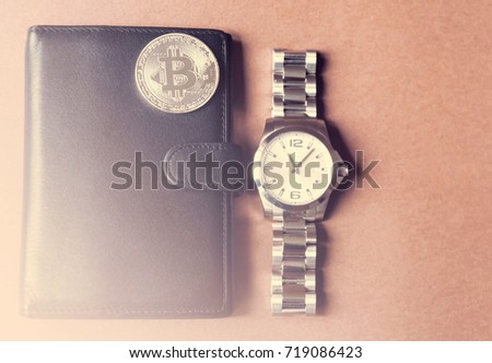 On a brown background is a black wallet, on it is a silver coin bitcoin, beside a man's watch. With a lighting.