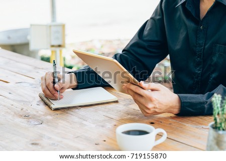 Business man hand holding tablet and writing notebook in coffee shop.