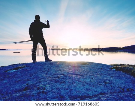 Rear silhouette of traveling man taking selfie at sea. Tourist with backpack standing on a rock, looking  over blue sea and taking photo by his smart phone camera