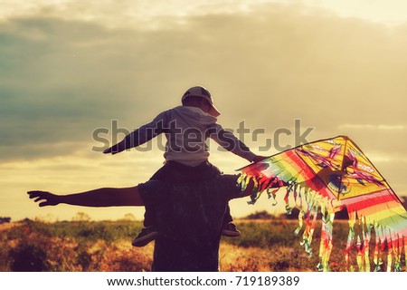 A boy with his father playing in a field with a kite .The child sits on the shoulders of the father