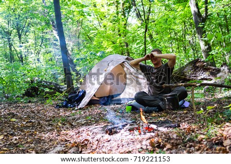 A bonfire and a tourist tent in the forest, the tourist is resting near the tent.