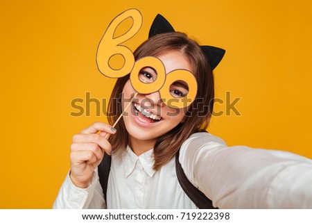 Close up portrait of a happy teenage schoolgirl in uniform with backpack taking a selfie while standing and holding paper mask isolated over orange background