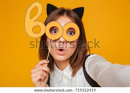 Close up portrait of a pretty teenage schoolgirl in uniform with backpack taking a selfie while standing and holding paper mask isolated over orange background