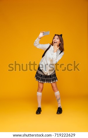 Full length portrait of a funny teenage schoolgirl in uniform with backpack taking a selfie while standing isolated over orange background
