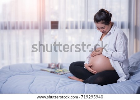 Young Asia pregnant, Pregnant wear white maternity clothes smile and look her tummy on bed with earphone and book
