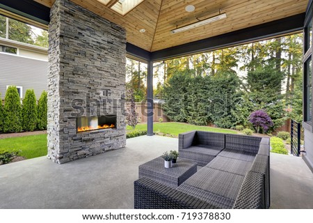 New modern home features a backyard with covered patio accented with stone fireplace, vaulted ceiling with skylights and furnished with gray wicker sofa placed on concrete floor. Northwest, USA 