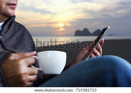 Mid adult man using smart phone to read latest online news or using app to connect with friends in social media and drinking black coffee on beautiful sea view beach in sunset time background.
