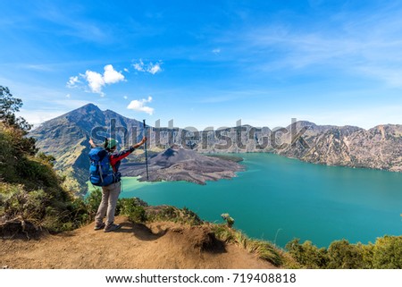 Hiker woman with blue backpack and trekking pole standing and enjoy with volcano Baru Jari Lake Segara Anak and Mount Rinjani summit view after finished climbing at Rinjani mountain. Lombok, Indonesia Royalty-Free Stock Photo #719408818