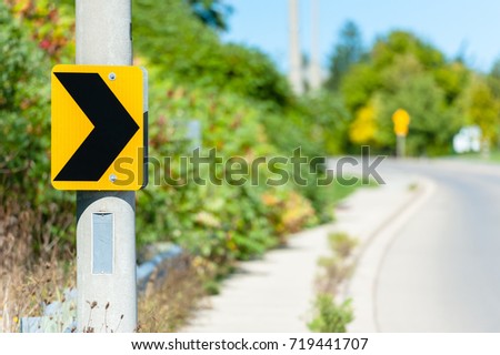 Black on yellow chevron road sign attached to post, indicating right turn against blurry curving road in background.