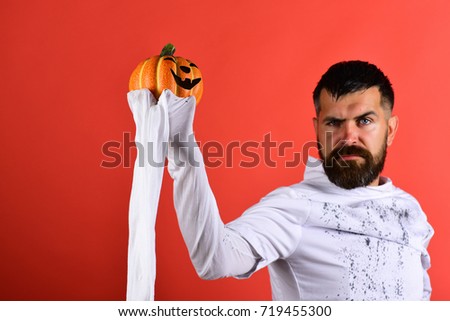 Man with serious face expression on red background. Halloween character in white long sleeved ghost costume. Guy with beard holds orange pumpkin with smile. Halloween, culture and traditions concept