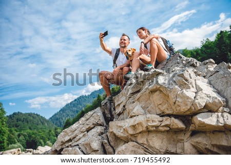 Hiker couple with small yellow dog sitting on mountain peak and  taking picture with smart phone
