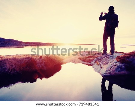 Rear silhouette of travelling man taking selfie at sea. Tourist with backpack standing on a rock, looking  over blue sea and taking photo by his smart phone camera