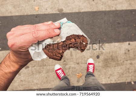 Young man standing on zebra crossing in the city with bitten Chocolate Cookie in his hand, point of view perspective. Royalty-Free Stock Photo #719564920