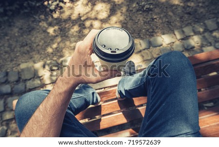 POV image of man sits on a bench and holding paper cup of coffee at a summer day in the park Royalty-Free Stock Photo #719572639