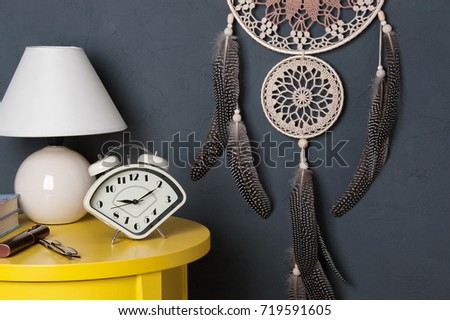 Beige brown crochet doily dream catcher, alarm clock and table lamp on yellow table in bedroom interior on gray background. Copy space for text.