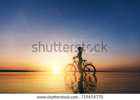The silhouette of a sporty girl in a suit that sits on a bicycle in the water at sunset on a warm summer day. Fitness concept. Sky background