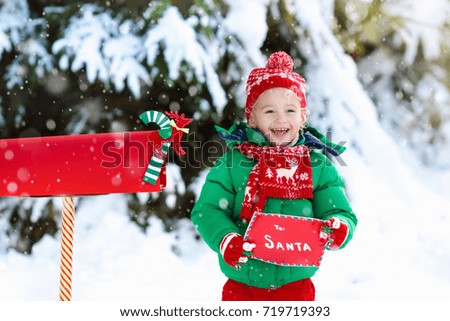 Happy child in knitted reindeer hat and scarf holding letter to Santa with Christmas presents wish list at red mail box in snow under Xmas tree in winter forest. Kids sending post to North Pole