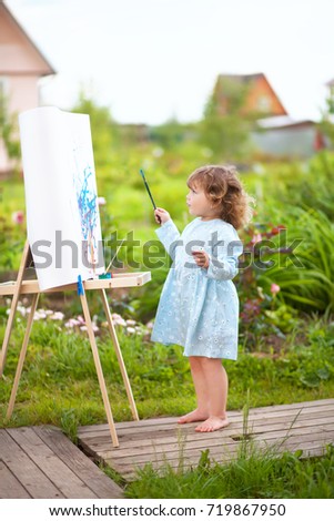 Adorable little painter. Cute toddler girl painting on the easel in the garden and having fun. Talented kids. Early art education.