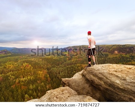 Hiker with knee joint stability bandage and forearm poles. Hiker on the cliff makes triumph gesture. Hilly forest landscape in background, nature park.