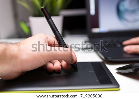 Closeup angled view shot of a retoucher working indoors using laptop computer and digital graphic tablet with a stylus pen. Green plant and an external hard drive aside on white office desk