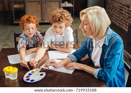 Painting our dreams. Selective focus on smart kids sitting next to their grandmother and choosing watercolor shades while holding painting brushes and creating new pictures.