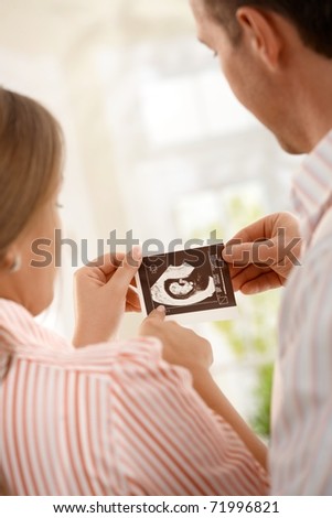 Expecting parents looking at ultrasound picture of baby together, mother pointing at photo at home. Focus on ultrasound image.?