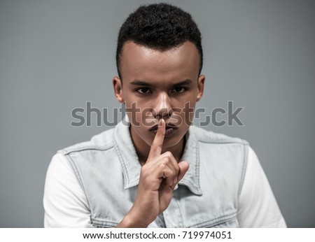 Attractive Afro American guy is showing silence sign and looking at camera, on gray background