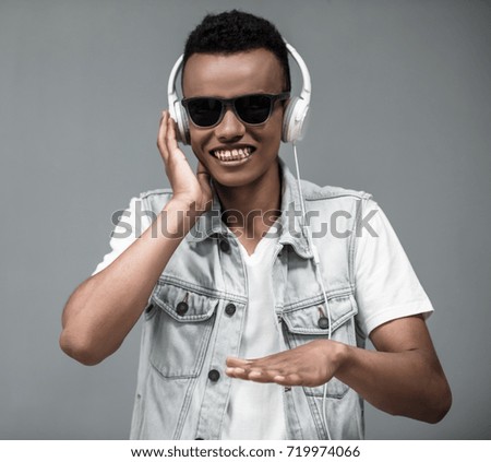 Stylish Afro American guy in headphones and sun glasses is listening to music and smiling, on gray background