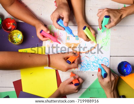 Hands hold colorful markers and draw kids illustration, top view. Artists wooden table with paints and colored paper. Markers in male and female hands draw on white paper. Creativity and art concept