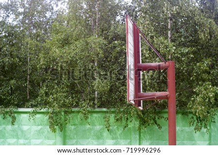 A street red basketball ring, rear view, against a background of green birch leaves and a green wall.