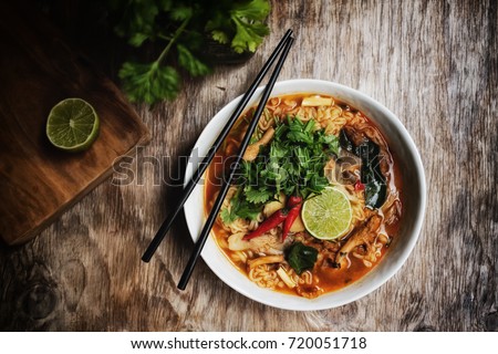 Ramen noodle with chanterelle mushroom in hot and spicy Thai style Tomyum soup. with lemon, chilis,coriander, lemon grass, kaffir or lime leaf and galangal. In ceramic bowl hand holding chopstick.  Royalty-Free Stock Photo #720051718