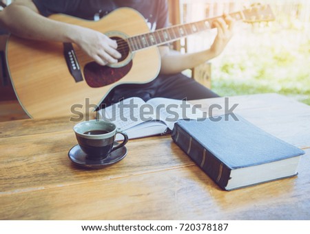 A young man is playing guitar and sing a song from Christian hymn  book with a cup of coffee and bible  on wooden table over garden background Royalty-Free Stock Photo #720378187