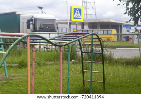 Summer playground, multi-colored pink and turquoise children's iron horizontal bar stairway among green grass, fence and road with lorries and bus, gray sky