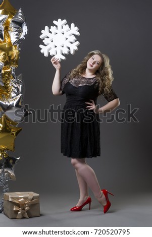 Plus size woman in black dres with gift box, snowflake and balloons on gray background, full length portrait