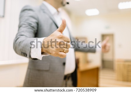 A successful business man showing thumbs up sign on the office background. Close-up of a confident entrepreneur in a suit.