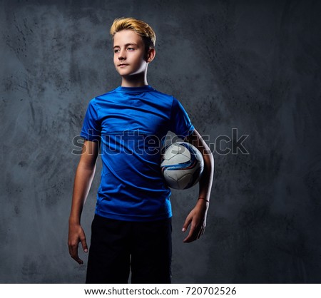 Blond teenager, soccer player dressed in a blue uniform holds a ball.