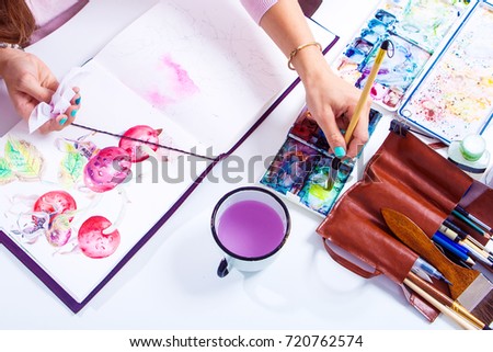 The dark-haired woman paints the pink flowers of peonies and wild rose with a wooden brush and watercolors in the drawing album, on the table lie a drawing bag, a leather case with brushes,,...