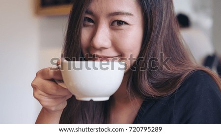 Closeup image of a beautiful Asian woman holding a white mug and drinking hot coffee with feeling happy in modern cafe 