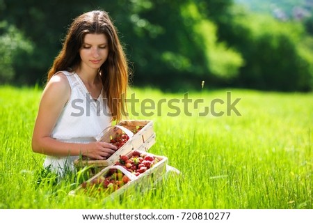 Beautiful brunette young girl sitting in the grass on a meadow with baskets full of strawberries. Place for your text. Healthy food and perfect summer sunshine day. Lovely picture.