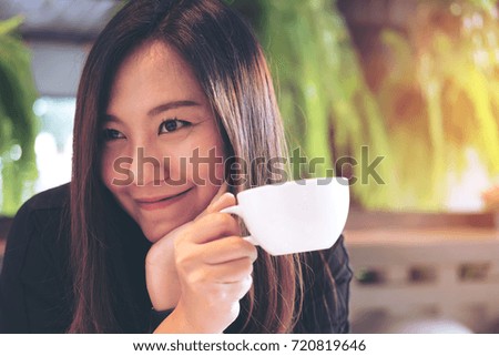 Closeup image of a beautiful Asian woman holding a white mug and drinking hot coffee with feeling happy in modern cafe and green nature background
