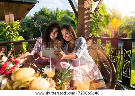 Two Young Women Use Digital Tablet While Breakfast On Terrace In Tropical Garden Beautiful Girls Hold Wireless Mobile Pc Sitting Together At Table Watch Photos In Morning