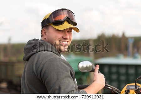 Caucasian man in sport protective goggles and yellow cap riding an ATV over rough terrain enjoying his adventure , smiling and showing thumbs up sign.